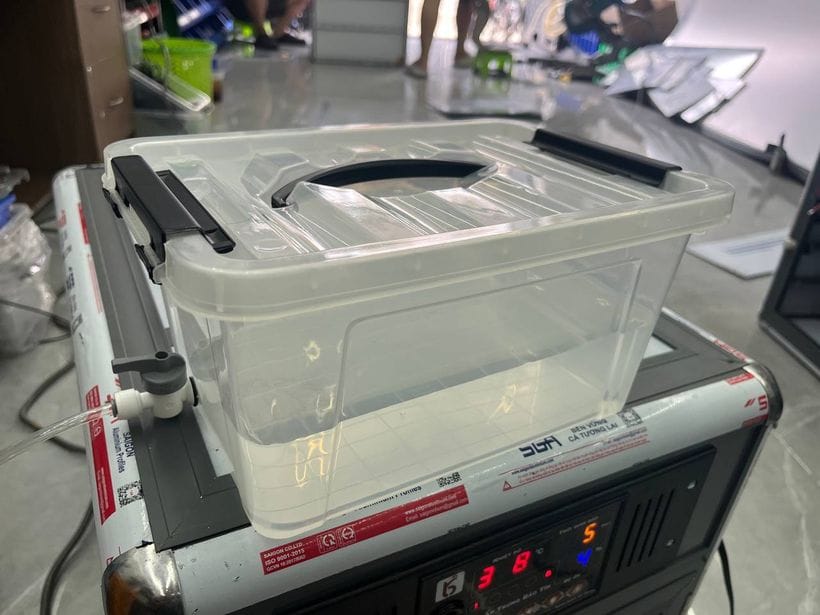 Bình cấp nước của máy ấp trứng Bảo Tín: hộp nhựa trong suốt có nắp gài và quai xách, đặt trên nóc máy, van khoá gắn ở thành bình nối với ống dẫn nước chạy xuống khay nước trong máy