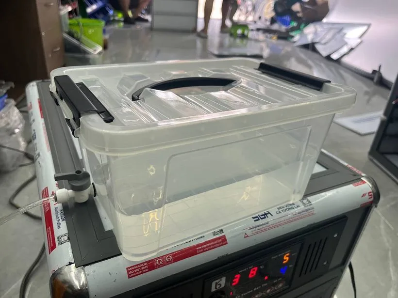 Bình cấp nước của máy ấp trứng Bảo Tín: hộp nhựa trong suốt có nắp gài và quai xách, đặt trên nóc máy, van khoá gắn ở thành bình nối với ống dẫn nước chạy xuống khay nước trong máy